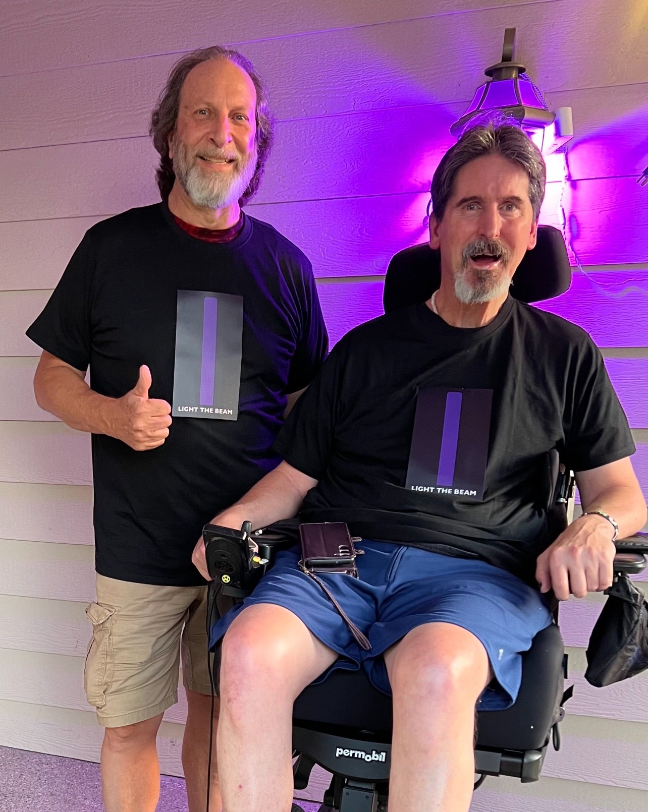 Two models side by side, one standing and one wheelchair user. Both wearing black t-shirt with panel that has a long purple beam in the middle of the chest with the words light the beam below it. Beam is turned on.