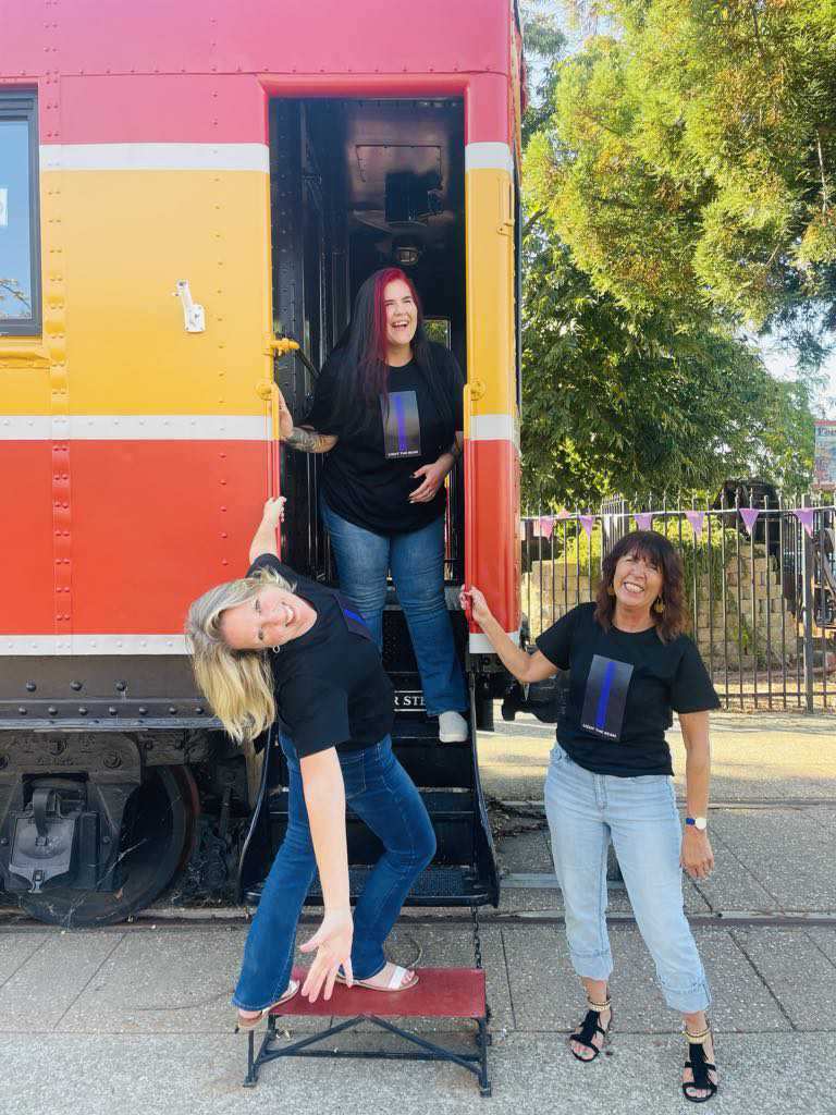 Picture of 3 female models smiling and laughing and having fun standing on entrance to a train painted yellow and orange. All 3 wearing black t-shirt with panel that has a long purple beam in the middle of the chest with the words light the beam below it.