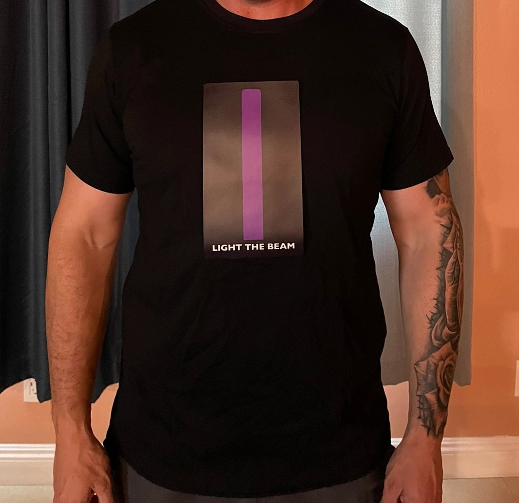 Picture of model from neck to waist, with tattoos on left arm, wearing black t-shirt with panel that has a long purple beam in the middle of the chest with the words light the beam below it. Beam is turned off.
