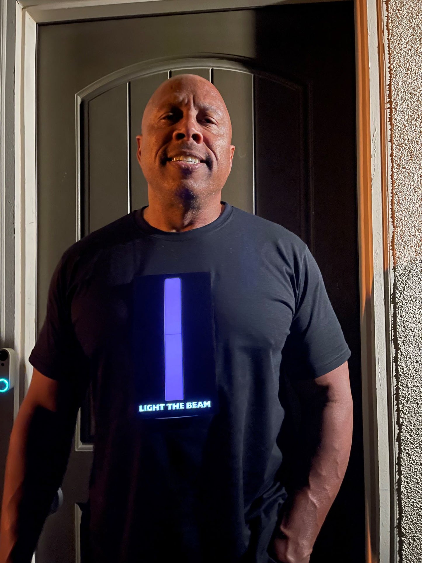 Picture of model standing in front of door. Model is
wearing black t-shirt with panel that has a long purple beam in the middle of the chest with the words light the beam below it.