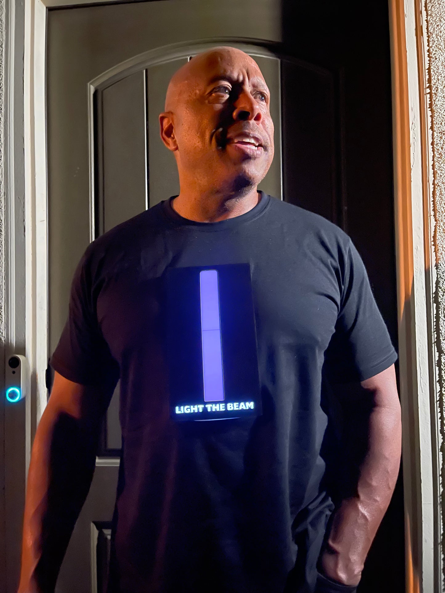 Picture of model standing in front of door looking to his lift. Model is
wearing black t-shirt with panel that has a long purple beam in the middle of the chest with the words light the beam below it.